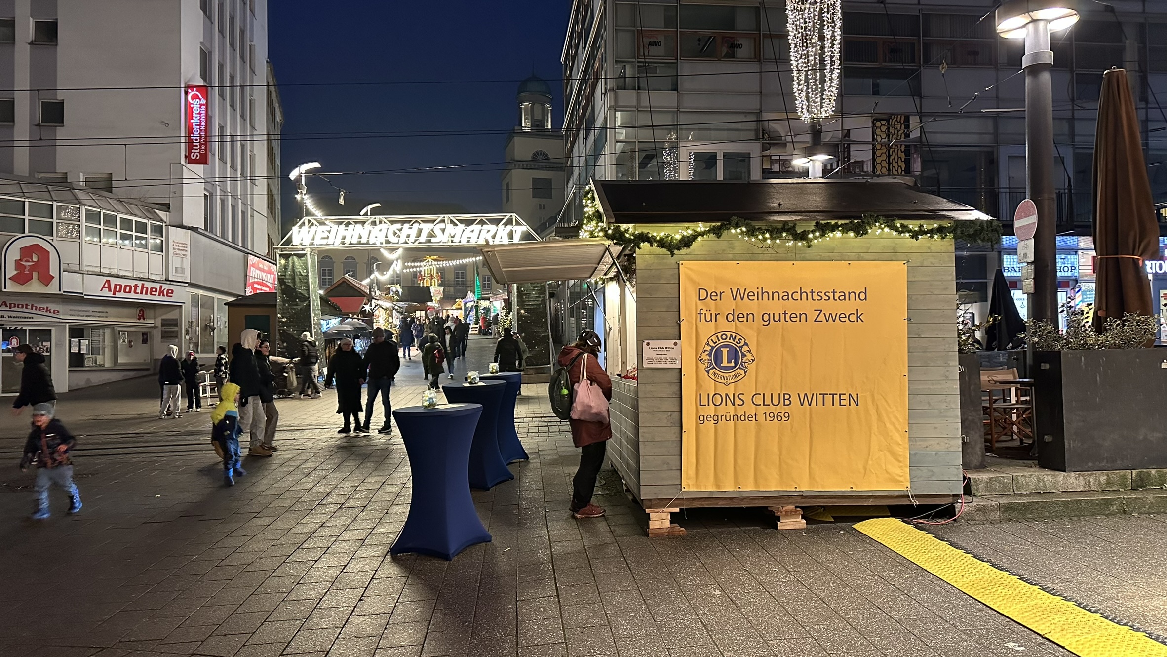 Beleuchteter Weihnachtsstand des Lions Club Witten mit gelbem Banner auf dem Wittener Weihnachtsmarkt 2024.