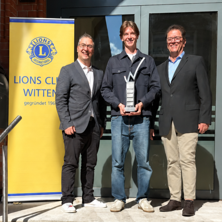 Drei Personen bei der Pokalübergabe neben einem Banner des Lions Club Witten; die Person in der Mitte hält den Wanderpokal.
